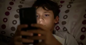 A boy lies in bed holding a smartphone above him.